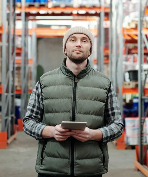 A warehouse worker holding a digital tablet, standing in a large inventory storage area with shelves filled with products, representing extensive inventory management